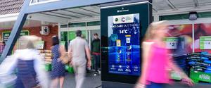 Shoppers passing in front of a digital display. The display shows an ad for Nivea sunscreen, with the day's current temperature included above the bottles.