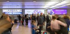 A large digital screen at Canberra airport. A crowd of people are walking by, collecting luggage from the conveyor belts.