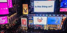 A busy New York City street with various billboards around. One looks like a giant Tweet with the words "Is this thing on?"